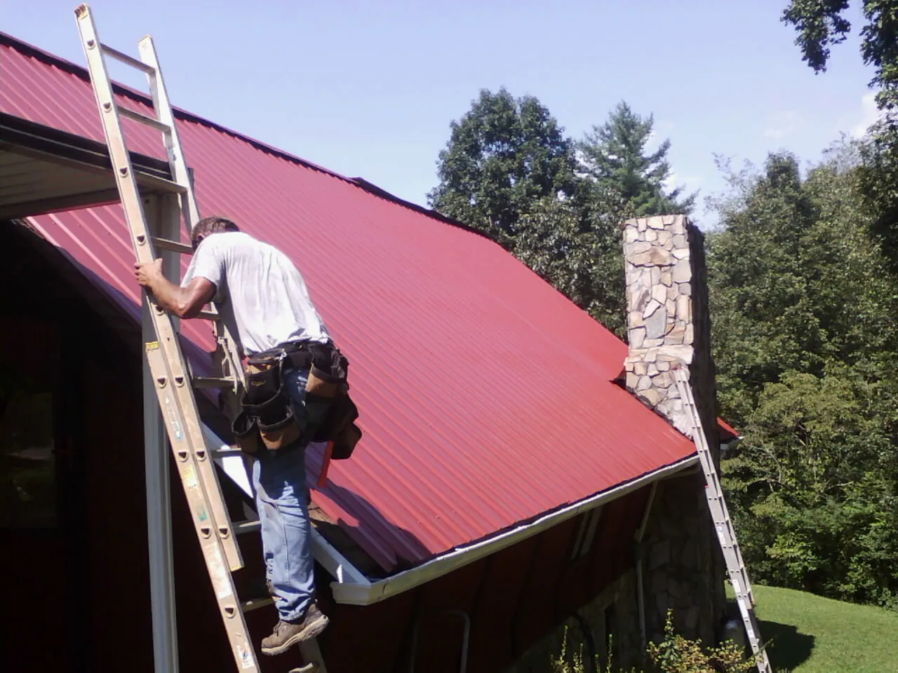 Roofer on a ladder beside a red metal roof and stone chimney — Soffit & Fascia Repair by Roofing Brothers in Russia
